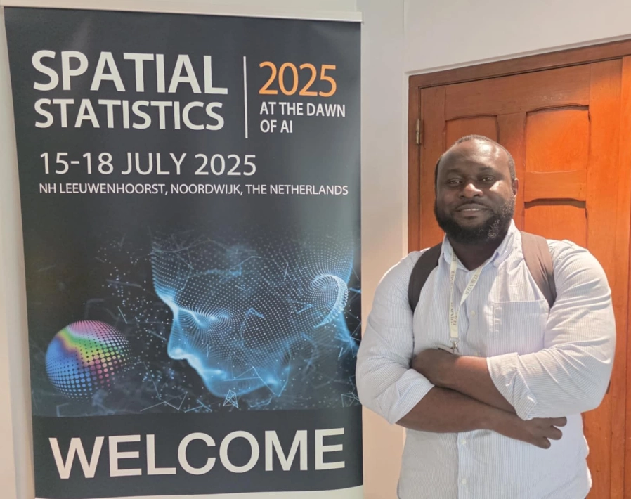 Photo of Dr Edson Utazi standing next to a Spatial Statistic 2025 conference poster