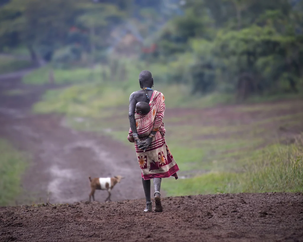 Photo of Suri Woman carrying a child while walking on a dirt road, Tulgit